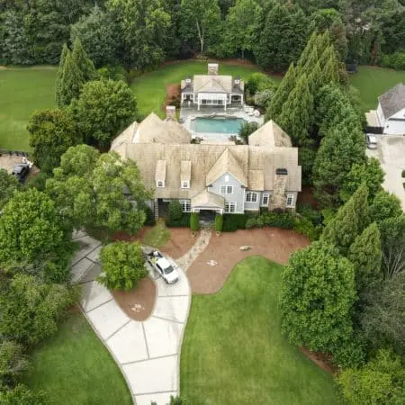 Drone image of upscale residential property with bright, freshly cleaned roof, circular driveway, and large backyard with pool surrounded by manicured trees