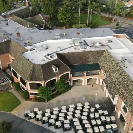 Drone photo shows large golf clubhouse with membrane and shingle roof cleaning in progress, rooftop equipment, and neatly arranged golf carts below