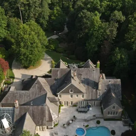Drone view of upscale property with darkened roof, large patio, and pool surrounded by forest, ideal candidate for soft wash roof restoration service