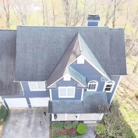 Aerial view of a blue two-story house with a gray shingled roof and white trim, with a paved driveway and manicured shrubs nearby.